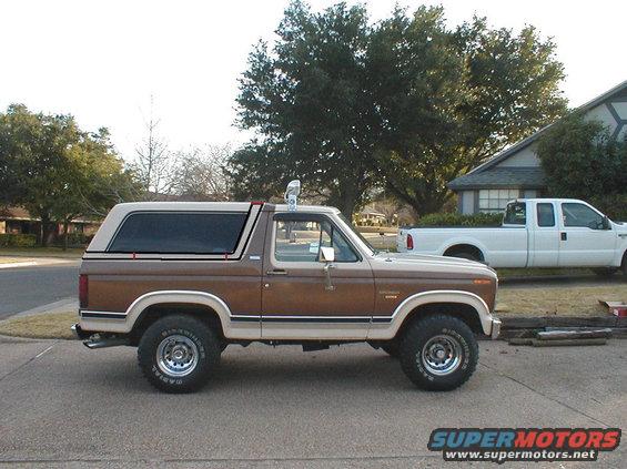 Brown and tan Ford Bronco with a camper shell, side view. Tires have white lettering