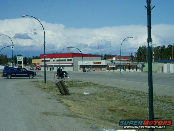 Motorcycle with rider on a road, blue pickup truck parked nearby, buildings in background