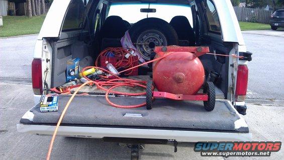 Red air compressor, hoses, and tools loaded in the bed of a white pickup truck. Spare tire visible in cab