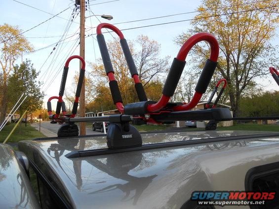 Thule kayak rack mounted on the roof of a light-colored SUV or truck, with a second vehicle visible in the background