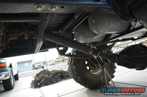 Underbody view of a vehicle's drive shaft and leaf springs, with a pickup truck visible on the left