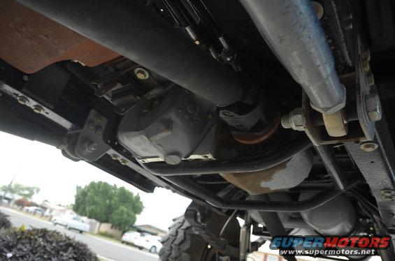Undercarriage view showing drive shaft, transfer case, and suspension parts. Other vehicles are visible in the background