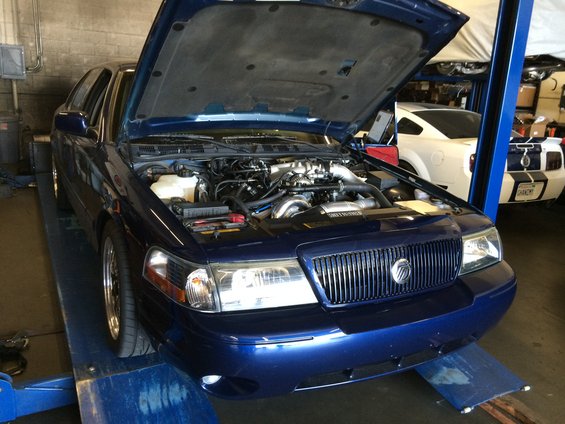 Blue 2003 Mercury Marauder on a lift with hood open, showing engine bay. Another car is visible in the background
