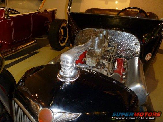 Black hot rod with chrome skull hood ornament and exposed V8 engine with dual carburetors