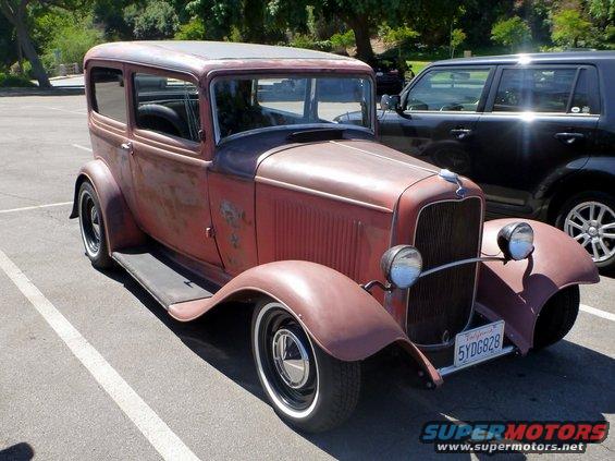 Rat rod sedan with rusty red paint and whitewall tires parked next to a black SUV. License plate reads 5YDGB28