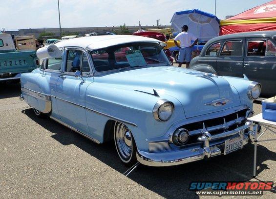 Light blue 1953 Chevrolet sedan with a white roof, lowered suspension, and chrome wheels. A teal pickup truck is visible to the left