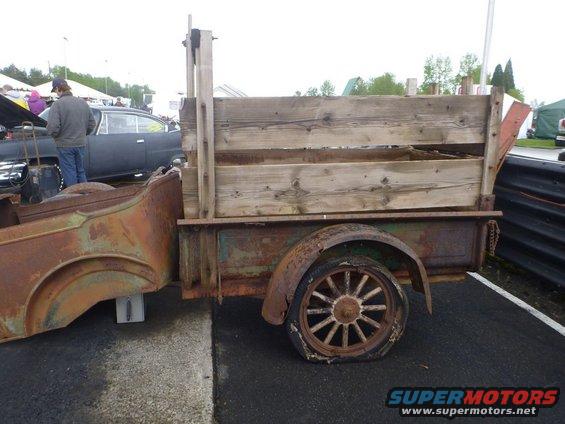 Rusty trailer with wooden bed and spoked wheels, flat tire. Another car is visible in the background