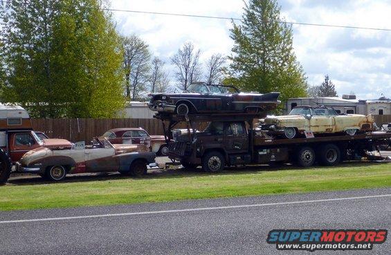 Several old cars, including convertibles, are loaded onto a car carrier. One car is on top of the carrier's cab