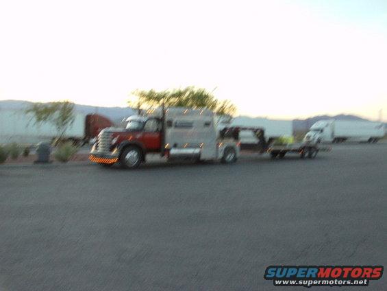 Tow truck with a flatbed trailer carrying a small vehicle, parked near other semi-trucks