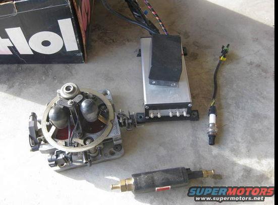 Holley carburetor, ignition box, and a spark plug on concrete. A black box with 'Hol' is in the background