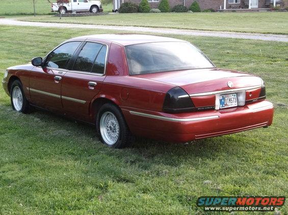 Red sedan parked on grass, rear 3/4 view. Blacked-out taillights, chrome trim, aftermarket wheels