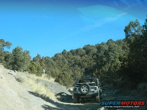 Silver Jeep Wrangler with spare tire on back driving on a rocky dirt road with trees and blue sky