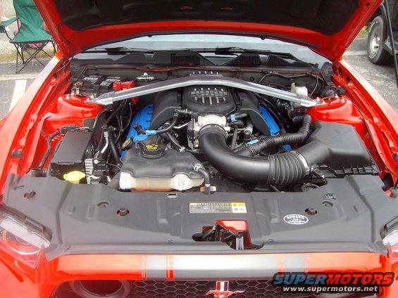 Red Ford Mustang engine bay with V8 engine, silver strut brace, and blue accents on valve covers. Supermotors.net watermark visible