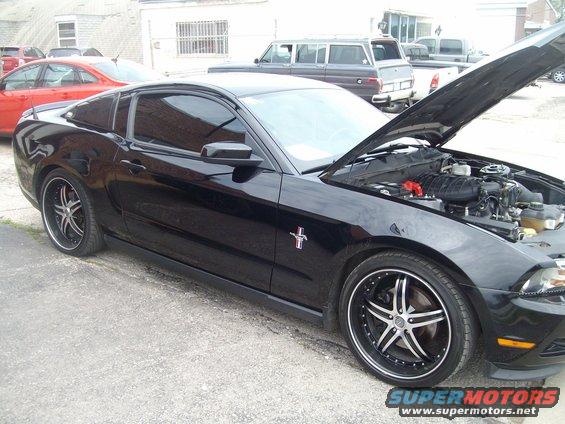Black Ford Mustang with hood open, showing engine and custom wheels. Red car in background