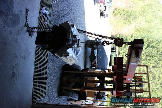 engine and transmission assembly suspended by a chain hoist over a forklift, with a dark SUV visible in the background