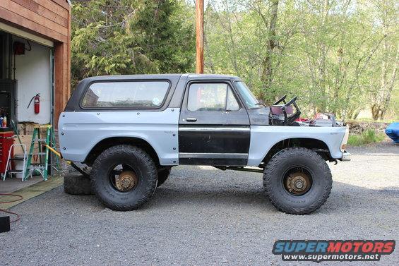 1978 Ford Bronco with primer gray body panels and black doors, lifted with large tires, parked outside a garage