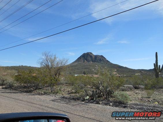 Desert scene with mountain, cactus, and power lines. Part of a car's side mirror is visible