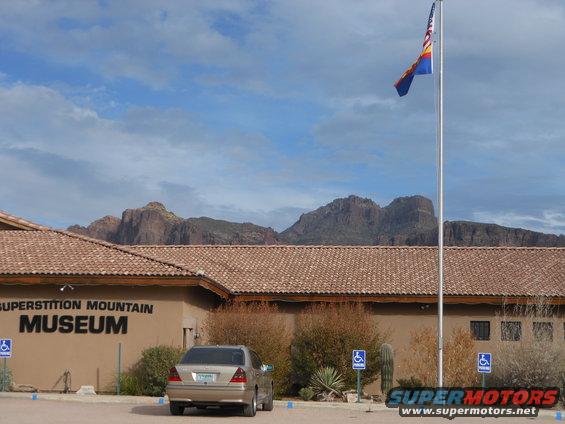 Superstition Mountain Museum building with mountains in background, Arizona flag flying, and a car parked out front