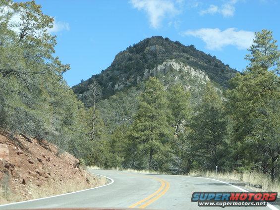 Winding road through a mountainous, forested area under a blue sky with clouds. Supermotors.net logo visible