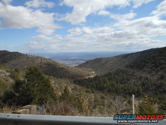 Mountain valley landscape with scattered trees and a cloudy sky, foreground has a metal guardrail