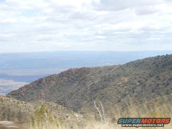 Mountainous terrain with dry grass in the foreground and a hazy valley with a town in the distance under a cloudy sky