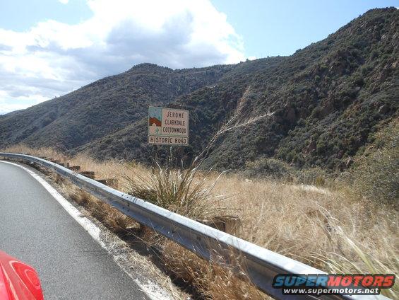 Red car fender in foreground, guardrail, dry grass, and a sign for Jerome, Clarkdale, Cottonwood Historic Road