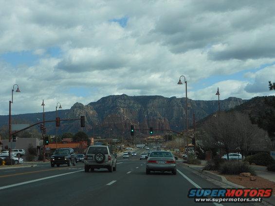 Driving on a road with traffic, passing a silver SUV and a black SUV, with large mountains in the background under a cloudy sky