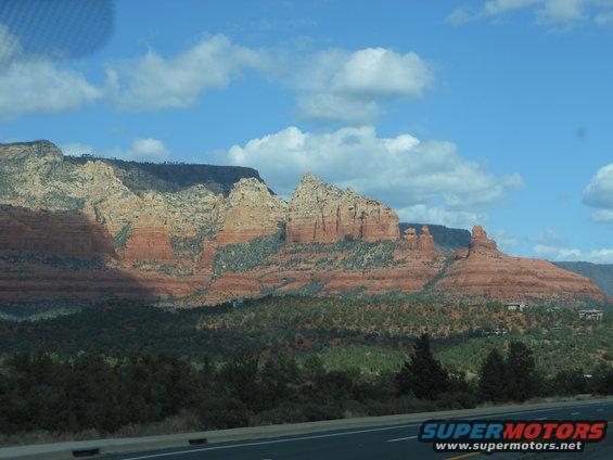Red rock formations and green trees under a blue sky with clouds, Supermotors.net logo visible