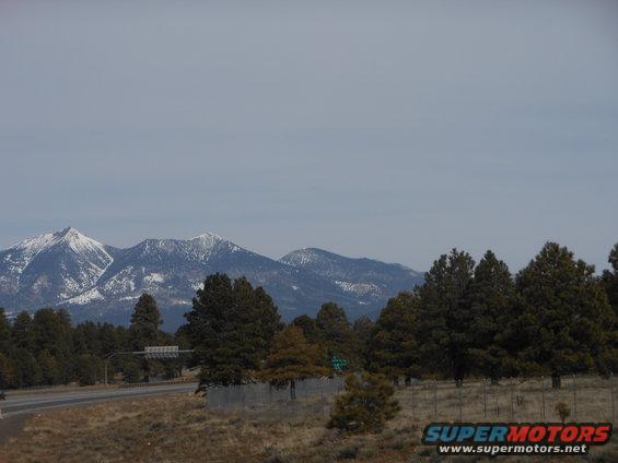 Mountain landscape with snow-capped peaks, pine trees, and a highway under a cloudy sky. Watermark reads SUPERMOTORS