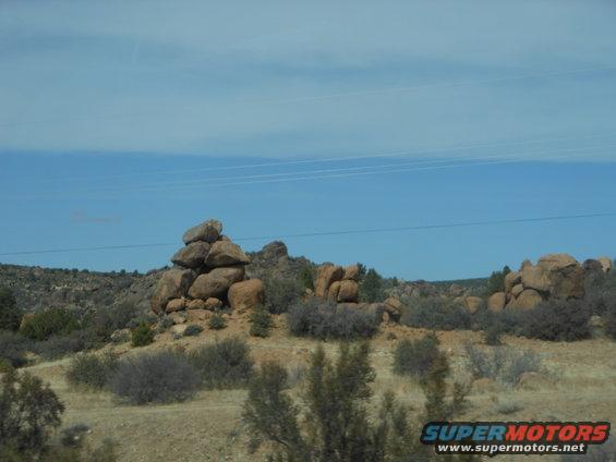 Rocky desert landscape with scrub brush and power lines against a blue sky. Supermotors.net logo in lower right