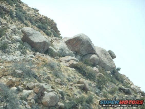 Rocky hillside with sparse vegetation under a bright sky. Watermark reads SUPERMOTORS