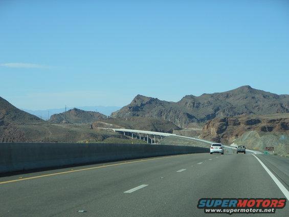 Two cars on a highway with mountains in the background. One white sedan and one silver SUV are visible