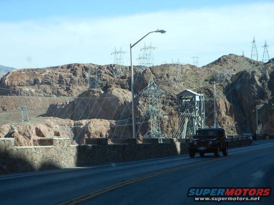 Red SUV driving on a road with a dam and power lines in the background