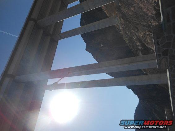 Looking up through concrete beams at a rocky cliff face and blue sky. Sun is bright