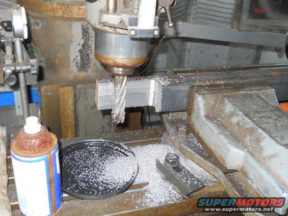 Milling machine cutting a metal block, with metal shavings scattered around. A can of spray is also visible