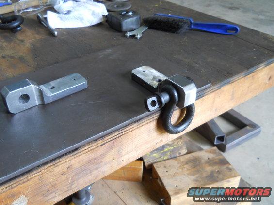 Two metal parts, one with a D-ring, sit on a workbench next to tools