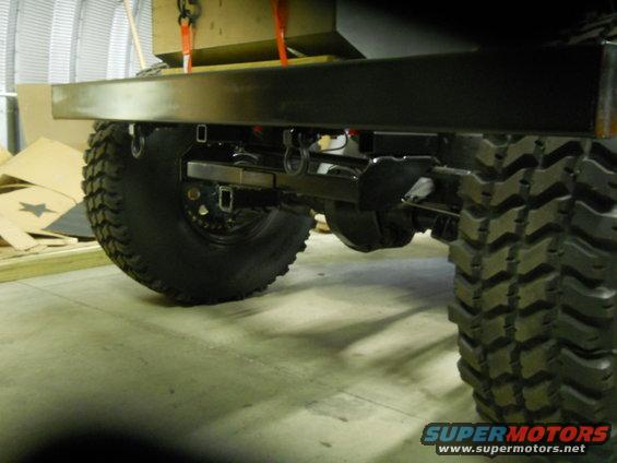 Rear axle, suspension, and large off-road tires of a vehicle. A metal bumper is visible above