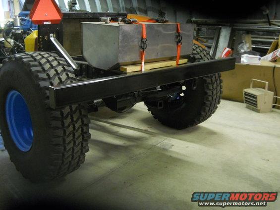 Truck chassis with large metal fuel tank strapped to the frame, large off-road tires with blue rims