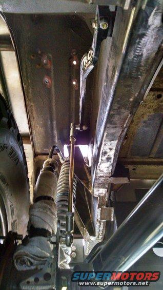 Underbody view of vehicle suspension, showing shock absorber, springs, and frame components. A 'TROS' logo is visible on a bracket