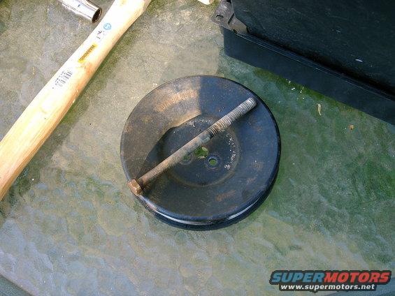 Smog pump pulley with a bolt through its center, resting on a glass table next to a hammer
