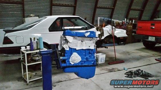 Blue engine on stand in garage, white car in background, red truck to the right. Supermotors.net logo visible