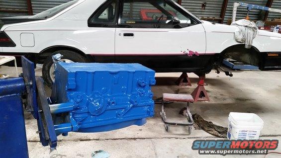White 1985 Ford EXP coupe on jack stands with a blue engine block in the foreground