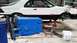1985 Ford Escort - A white 1985 Ford EXP coupe is elevated on jack stands, with a freshly painted blue engine block positioned in front of it.