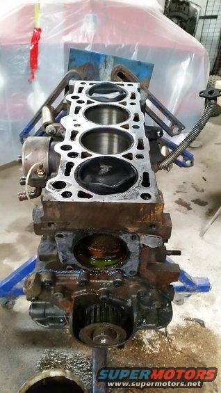 Engine block on a stand, showing cylinders and some internal components. Text at bottom reads SUPERMOTORS.NET