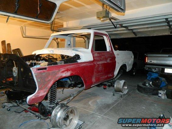 1979 Ford F-150 truck body in red and white primer, undergoing restoration with front end disassembled. Another truck is visible in the background