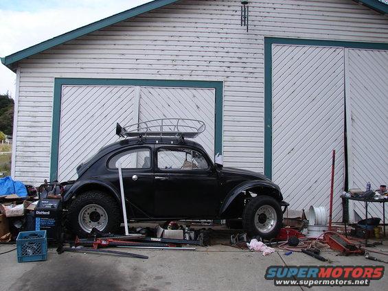 Black 1964 Volkswagen Beetle with a roof rack, modified suspension, and off-road tires parked in front of a garage