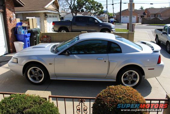 Silver coupe with a hood scoop and spoiler, parked in a driveway. A dark pickup truck is behind it