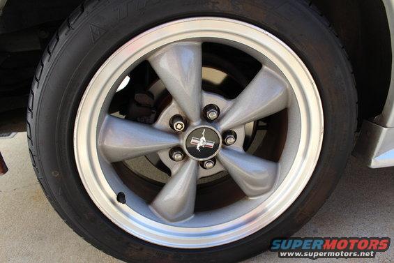 Close-up of a silver 5-spoke wheel with a black center cap a running horse emblem. Tire is black with visible tread