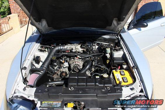 Silver 2002 Ford Mustang engine bay with hood open, showing V8 engine, aftermarket intake, and yellow battery