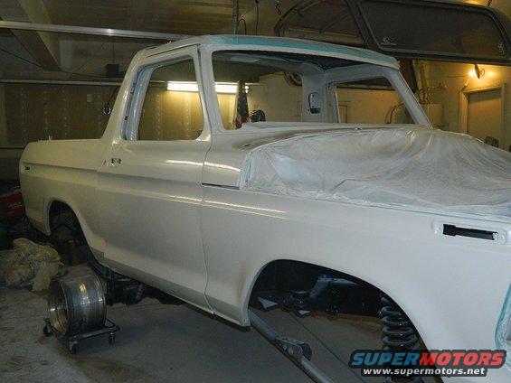 White 1978 Ford Bronco body, partially covered in plastic, in a workshop. Front fender and door visible
